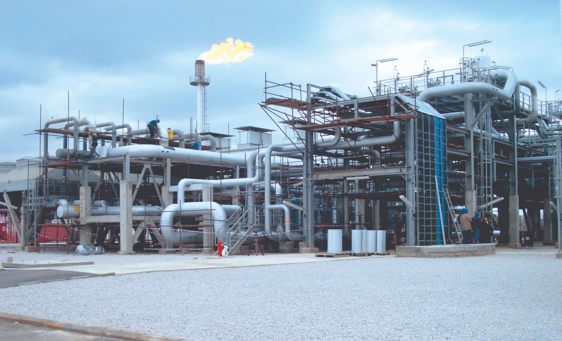 Industrial gas processing facility with workers on scaffolding and interconnected piping; gas flare burning in the background under a cloudy sky