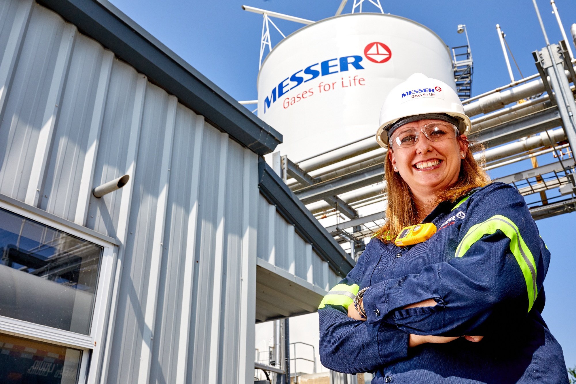 Smiling Messer employee in safety gear stands in front of an industrial gas facility with a large storage tank labeled “Messer – Gases for Life”
