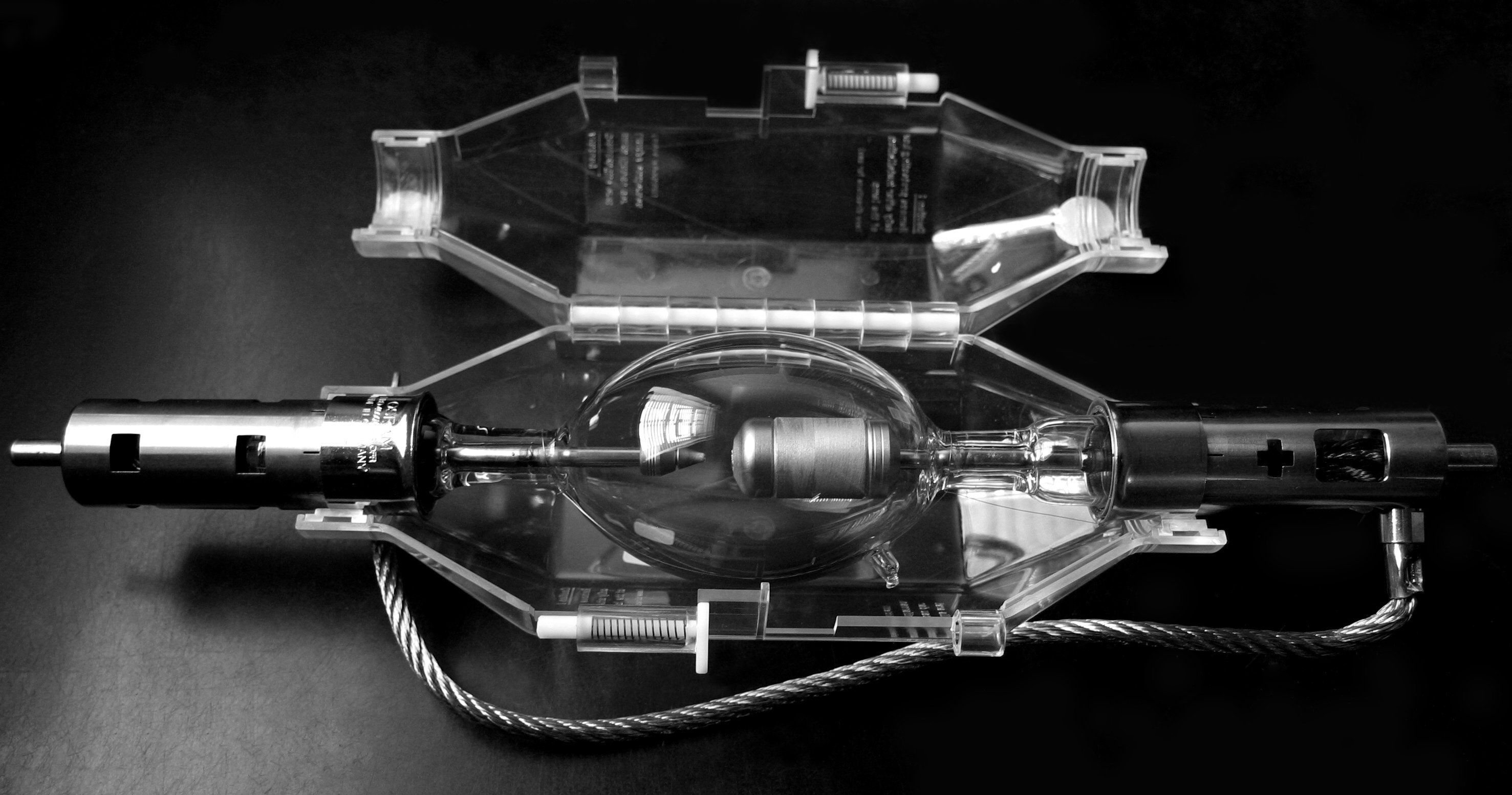 A high-intensity xenon arc lamp in a clear protective case placed on a dark surface. The lamp’s central glass bulb, metal electrodes, and wiring are visible, along with latches and molded plastic components of the case.